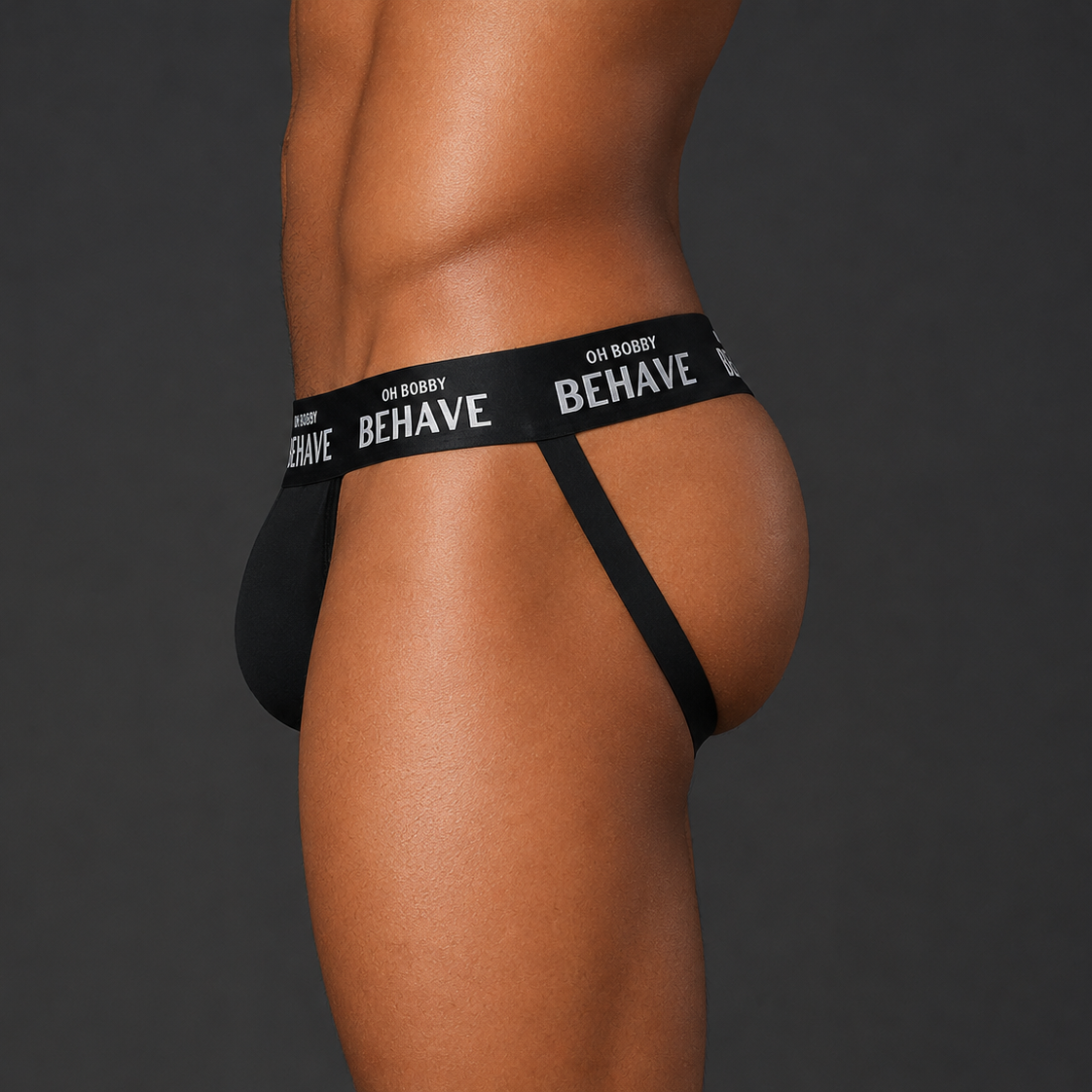 A close-up side view of a model wearing black thong underwear with a wide black waistband printed with the white text 'BEHAVE' and 'ON BOY'.