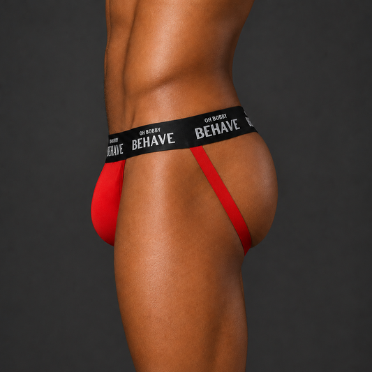 A close-up side view of a model wearing a red jockstrap underwear bottom with a black waistband printed with the brand name OH BOBBY BEHAVE.